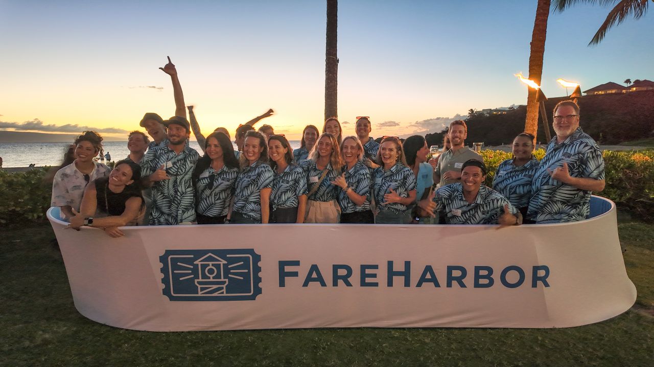 FareHarbor team at a Hawaiian beach meetup at sunset, standing behind a branded banner